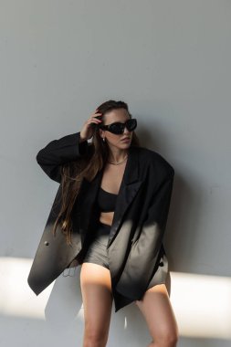 Makyajı az, spor ceketi çok büyük ve güneş gözlüklü zarif bir kadın doğal güneş ışığında şık bir şekilde poz veriyor..