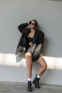 Büyük boy siyah ceketli şık bir genç kadın gri bir duvara karşı poz vermiş, modern sokak modasını ve doğal ışık altında kendine güvenen tavırlarını sergiliyor..