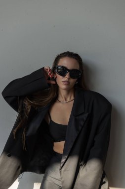 Şık, büyük spor ceketli ve fütüristik güneş gözlüklü genç bir kadın, güneş ışığında dışarıda poz veriyor, sokak modası için mükemmel..