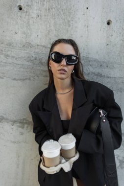 Şık bir şehir. Büyük spor ceketli, güneş gözlüklü, elinde kahve taşıyan bir kadın.