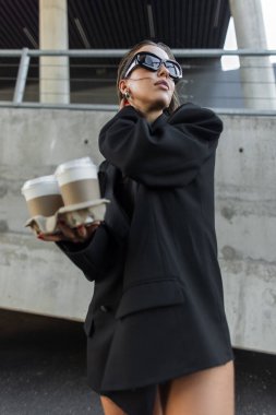 İki paket kahve fincanı tutan şık siyah ceket ve modern güneş gözlüğü takan modaya uygun genç bir kadın. Kahveli zarif bir genç kadın, büyük beden ceket ve şık güneş gözlüğü takıyor..