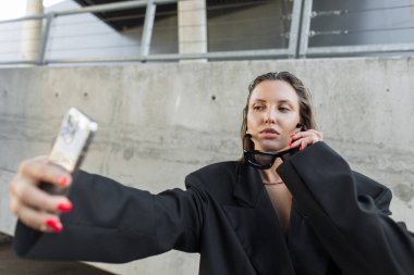 Kendine güvenen, büyük spor ceketli ve koyu güneş gözlüklü bir model. Selfie için akıllı telefon tutuyor. Zarafeti, cesur stili ve şehir havasını temsil ediyor..