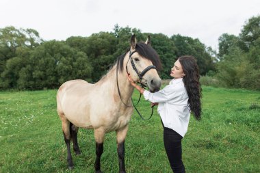 Beyaz gömlekli güzel bir kadın yeşil alanda atıyla yürüyor. At dostluğu ve özen. Kırsal yaşam