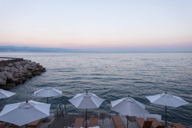 Gün batımında Adriyatik Denizi kıyısında şemsiyeler ve güneşliklerle Hırvatistan 'ın huzurlu deniz manzarası. Hırvatistan 'ın deniz manzarası, gün batımında beyaz şemsiyeler ve tahta güneşliklerle sakin Adriyatik manzarası