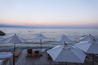 Hırvatistan 'da gün batımında beyaz şemsiyeler ve tahta güneşliklerle deniz manzarası, sakin Adriyatik Denizi ve uzak dağlara bakıyor. Mükemmel bir yaz tatili ve seyahat geçmişi