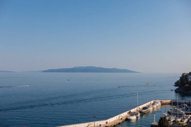 Hırvatistan 'ın Opatija kentindeki sakin Adriyatik kıyıları, güneşli bir günde açık deniz ve uzak bir ada zeminine karşı marina, iskele ve demirlemiş teknelerle dolu..