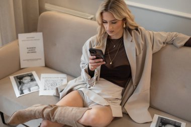 Şık giyinmiş, ceketli, güzel, sarışın bir kadın kanepeye oturmuş, akıllı telefonuna bakıyor. Profesyonel iş kadını çalışıyor.