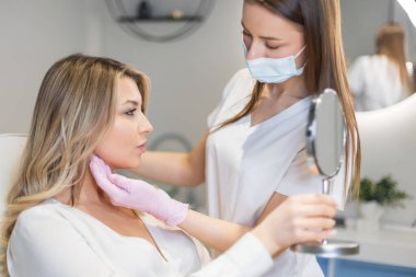 Kozmetik uzmanının randevusundaki güzel bir kadın ve güzellik kliniğindeki aynaya bakıyor. Doktor kozmetik uzmanı prosedürleri yapar.