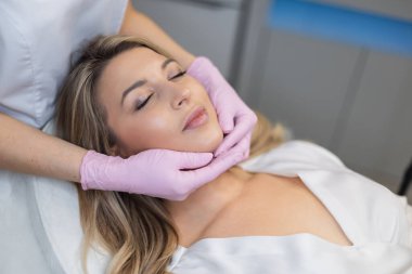 Güzellik kliniğinde temiz cilt ve dudakları olan güzel bir genç kadın. Pembe eldivenli doktor kozmetik uzmanı ameliyatlar ve yüz masajı yapar.