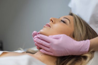 Güzellik kliniğinde temiz cilt ve dudakları olan güzel bir genç kadın. Pembe eldivenli doktor kozmetik uzmanı ameliyatlar ve yüz masajı yapar.