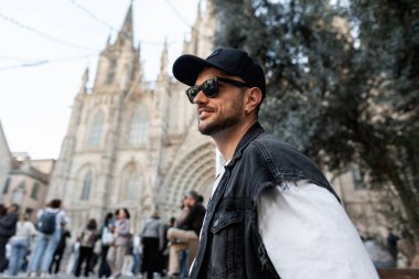 Siyah şapkalı, güneş gözlüklü, beyaz tişörtlü, kot yelekli yakışıklı adam Barcelona 'da klasik kilisenin yanında yürüyor.