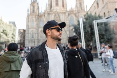 Güneş gözlüklü, siyah şapkalı, beyaz tişörtlü, kot yelekli yakışıklı hippi Barselona 'da kilisenin yanında yürüyor.