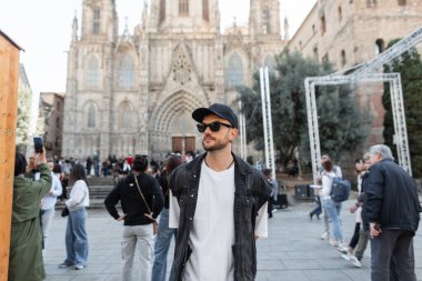 Siyah şapkalı, modaya uygun giyinen, modaya uygun giyinen, gözlüklü yakışıklı bir adam kilise yakınlarında Barcelona 'da yürüyor. İspanya 'ya seyahat