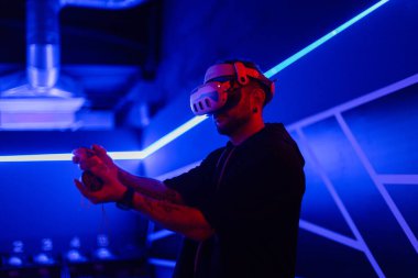VR gözlüklü yakışıklı havalı oyuncu elinde joystickle neon ışıklı oyun odasında sanal oyun oynuyor. Sanal gerçeklik teknolojisi