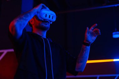 VR kulaklıklı havalı teknik adam karanlık odada neon ışıklı sanal menüyü kontrol ediyor.