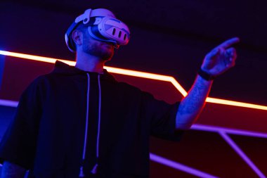 VR kulaklıklı havalı teknik adam sanal menüyü kontrol ediyor ve karanlık odada neon ışıklı parmaklarla dokunuyor.
