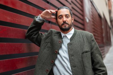 Yakışıklı hippi stili sakallı ve bıyıklı moda kıyafetleriyle Barcelona şehrinin kırmızı duvarının yanında duran adam.