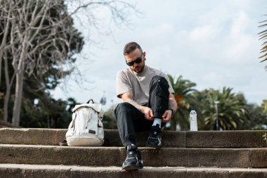 Yakışıklı hippi stil gezgini, güneş gözlüklü, moda elbiseli, spor ayakkabılı, sırt çantalı ve su dolu Barcelona 'da parkta merdivenlerde oturan.