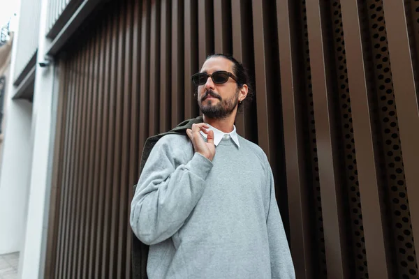 Yakışıklı, sakallı, bıyıklı, güneş gözlüklü, şık giyinmiş, şehirde bronz metal duvarın yanında yürüyen bir moda adamı.