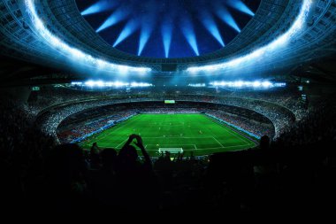 Geceleri yıldızlı modern futbol stadyumu. Mavi projektörler yeşil perdeyi aydınlatıyor ve yıldızlı gökyüzünün altında kalabalığı alkışlıyor. Spor enerjisi. Taraftarlarla futbol maçı. Dünya Kupası Şampiyonları. Kavram