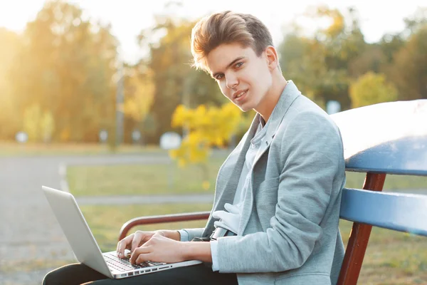 Pohledný mladík sedící na lavičce v parku při západu slunce. Drží notebook v rukou a pracovní.. — Stock fotografie