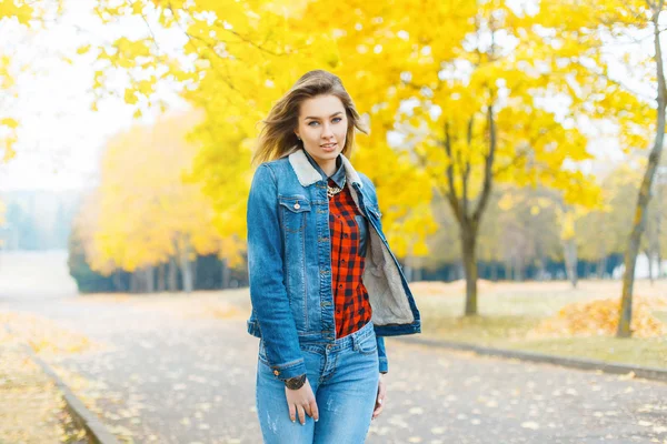 Menina bonita em roupas de jeans e uma camisa quadriculada vermelha no parque de outono . — Fotografia de Stock