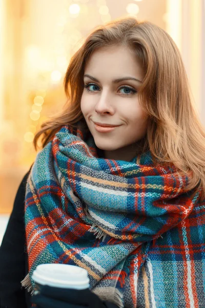 Young beautiful girl in stylish knitted scarf and black coat drinking coffee on the background of shop windows — Stockfoto