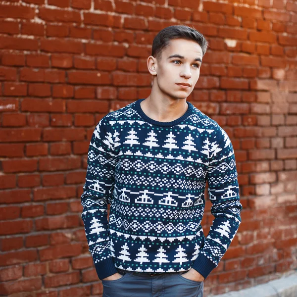 Jovem homem bonito em uma camisola de malha com ornamentos de Natal em um fundo de parede de tijolo vermelho — Fotografia de Stock