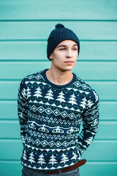 Bonito jovem em um suéter de Natal de malha e chapéu contra um fundo de parede de madeira azul . — Fotografia de Stock