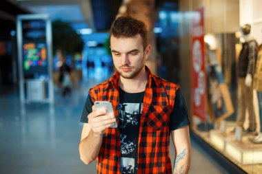 Young handsome man in the red shirt is holding a phone on the background of a shopping center