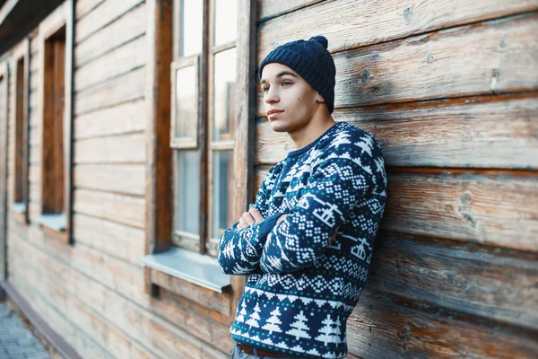 Handsome young man in a white knit sweater and cap near a wooden house. — Stockfoto