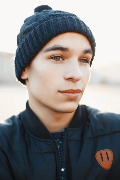 Retrato de close-up de um jovem em um chapéu de malha quente e jaqueta de inverno em um fundo de um pôr do sol — Fotografia de Stock