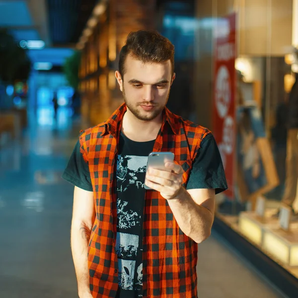 Young handsome guy in the red shirt is holding a phone on the background of a shopping center with a showcase — Stock fotografie