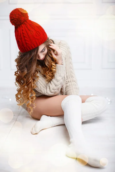 Beautiful fashionable American girl in knitted warm clothes on a background of lights. Woman sitting on white wooden floor — Stock Photo, Image