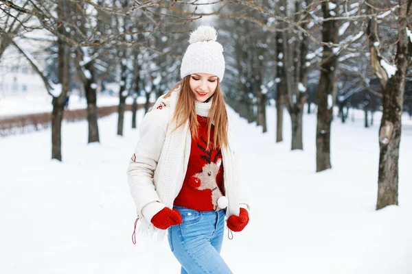 Bella donna con un maglione rosso, un berretto bianco e una giacca, sciarpa lavorata a maglia cammina vicino a un albero nel parco — Foto Stock