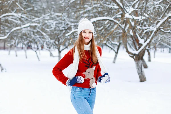 Menina elegante bonita em um boné de malha, suéter vermelho com um veado e em um fundo branco nevado com árvores . — Fotografia de Stock