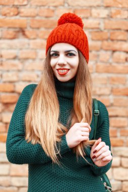 Beautiful cute girl in a red cap and green knitted jacket standing near red brick wall