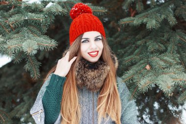 Young beautiful girl with red lips and a knitted hat in winter coat standing near tree