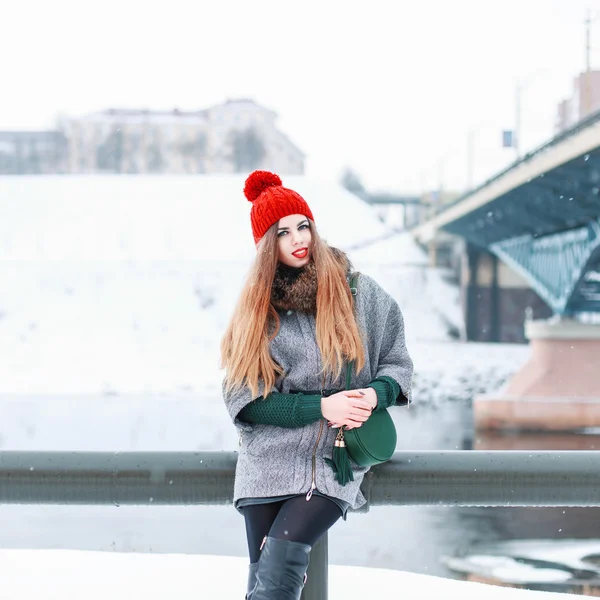 Young beautiful girl in a fur coat and a knitted cap sits on a background of the bridge on a winter day — Stok fotoğraf