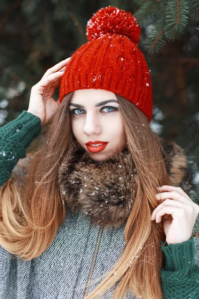 Pretty girl with beautiful blue eyes in a red hat and coat standing near spruce — Stock Fotó