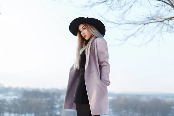 Joven chica con estilo en un sombrero y abrigo se encuentra en una colina sobre un fondo de un paisaje de invierno —  Fotos de Stock