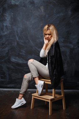 Pretty woman with blonde hair in a stylish black shirt, white sneakers and torn jeans sitting on a wooden chair on a dark background with chalk stains