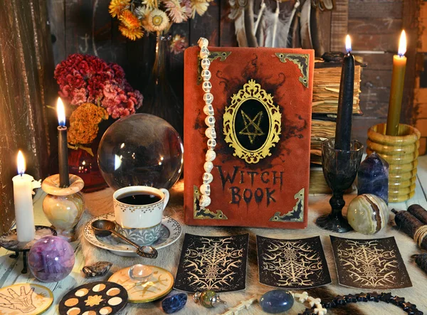 Red witch book with tarot cards, tea cup, black candle and magic ritual objects on the table. Esoteric, gothic and occult background, Halloween mystic concept.