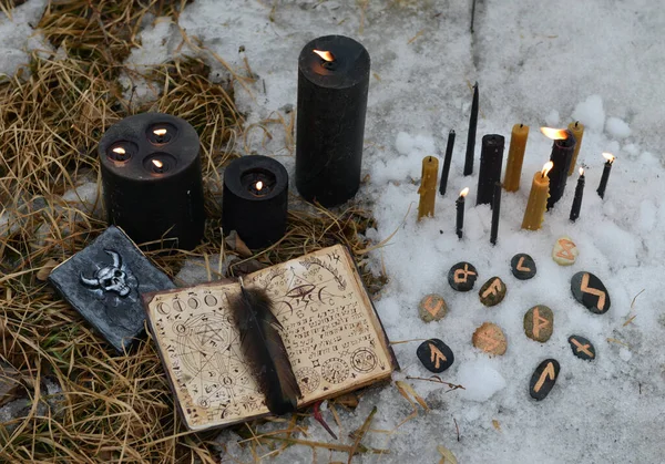 Witch ritual of black magic with open book of spell, runes and candles in snow. Esoteric, gothic and occult background, Halloween mystic and wicca concept outdoors.