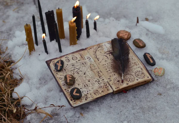 Open with book of magic spells, runes on stones and candles in snow. Esoteric, gothic and occult background, Halloween mystic and wicca concept outdoors.