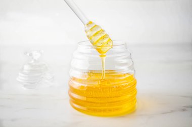 Bal, bal kaşığından cam kavanoza akar. Şeffaf bir kavanozun içindeki sağlıklı, organik, kalın bal beyaz bir masada duruyor. Yakın plan.