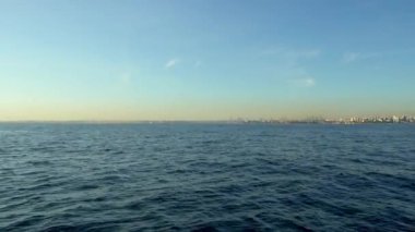 Mavi denizin insansız hava aracı görüntüsü. Şehir ufukta görünür, ufuktaki gökyüzü soluk sarı, güzel bir gündoğumu. İnsansız hava aracı ilerliyor.