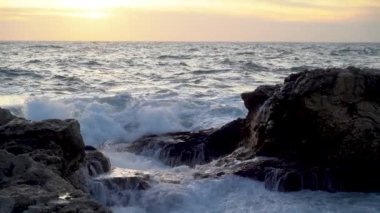 Dalgalı denizin büyüleyici bir görüntüsü. Dalga güçlü bir şekilde ön plandaki kıyı kayalarının üzerinden geçiyor ve ona karşı kırılıyor. Büyük bir sprey ve köpük duvarı yükselir. Ufukta, deniz gün batımında altın bir gökyüzünde kaybolur. Yavaş çekim.