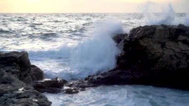 Dalgalı deniz ve kıyı kayalarının nefes kesici yakın görüntüsü. Dalgalar su yüzeyinde yuvarlanır, güçlü bir şekilde kayalara çarpar, beyaz köpük ve spreyden kolonlar oluşturur. Deniz gün batımında kırmızı ve altın gökyüzü ile buluşur. Yavaş çekim.