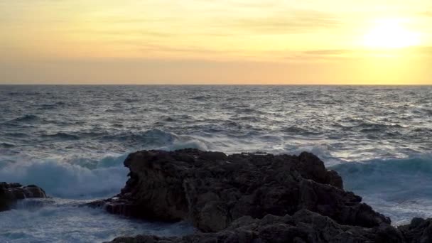 日没時の穏やかな海の景色 波は美しく力強く海岸の石の上を転がり それらに対して衝突する スプラッシュ立ち上がりと白い泡は水の上にパターンを作成します 前方には美しいオレンジ色の空と明るい夕日です — ストック動画